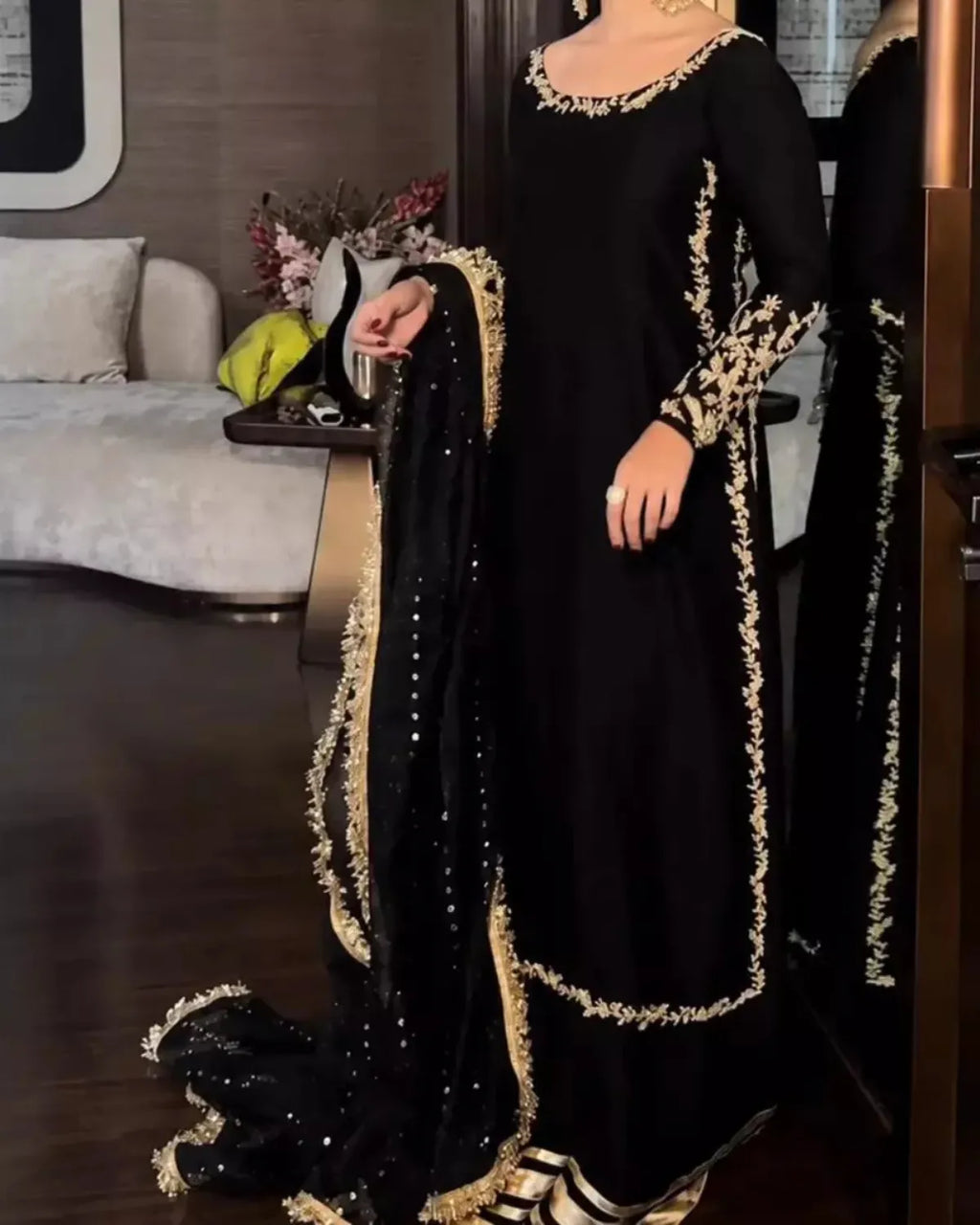 A woman wearing a black traditional outfit with ornate gold embroidery and a matching black dupatta with gold trim, standing in an indoor room with a mirror behind her.
