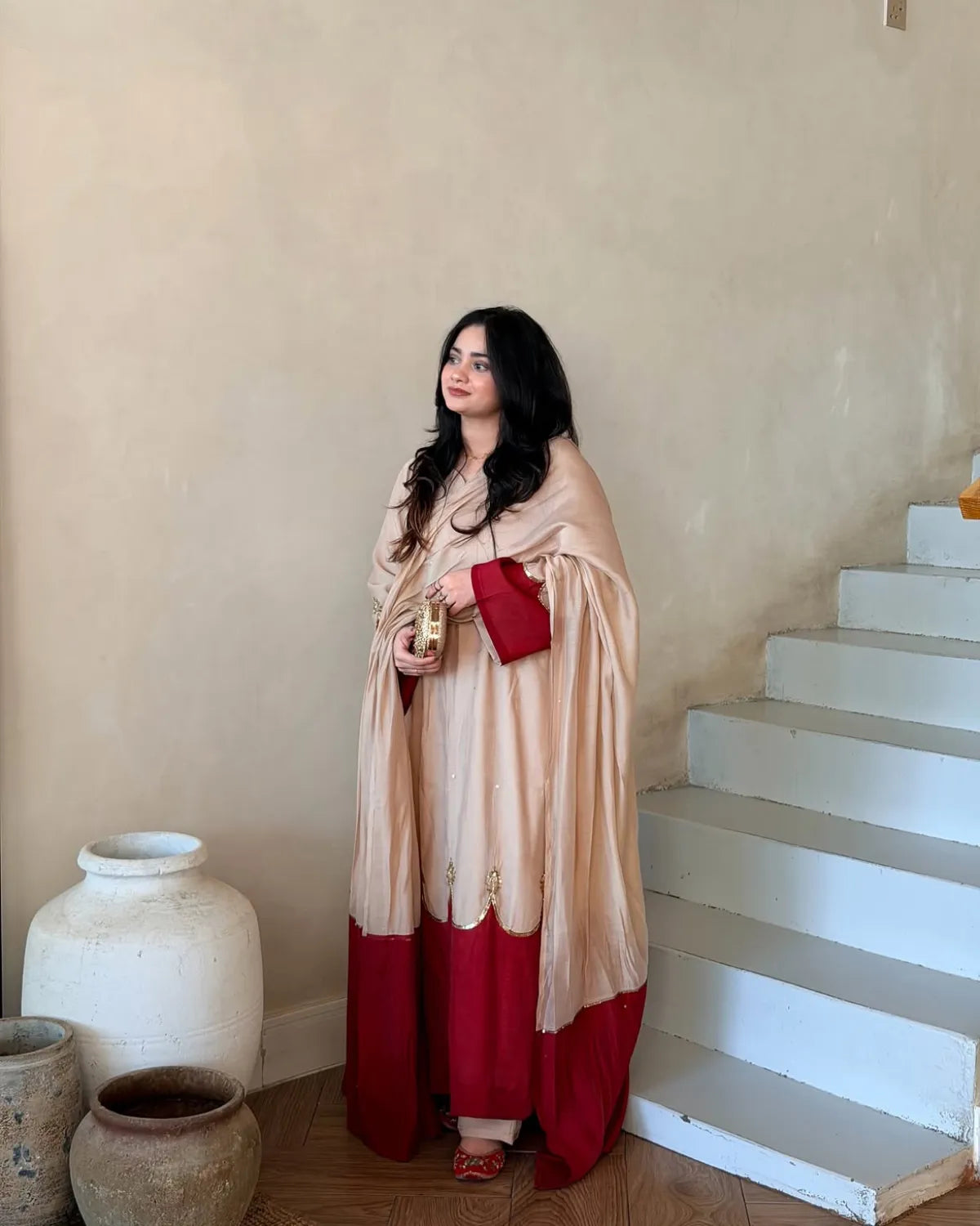 A woman stands indoors near a staircase and large ceramic pots, wearing a long beige and red traditional outfit with a draped shawl and holding a red clutch.