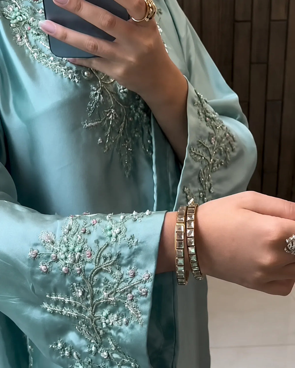 A close-up of a person wearing a light blue-green satin garment with intricate silver and pink floral embroidery on the sleeves, holding a phone in front of their face.