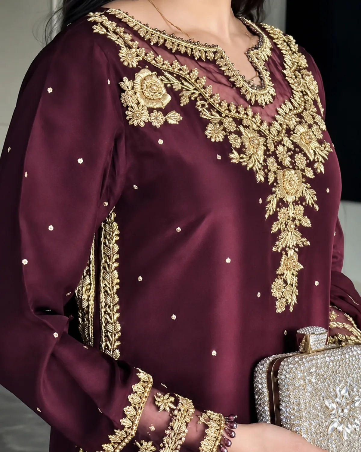 A close-up of a woman wearing a deep maroon long-sleeve kurta with ornate gold floral embroidery and small silver embellishments, holding a silver clutch.