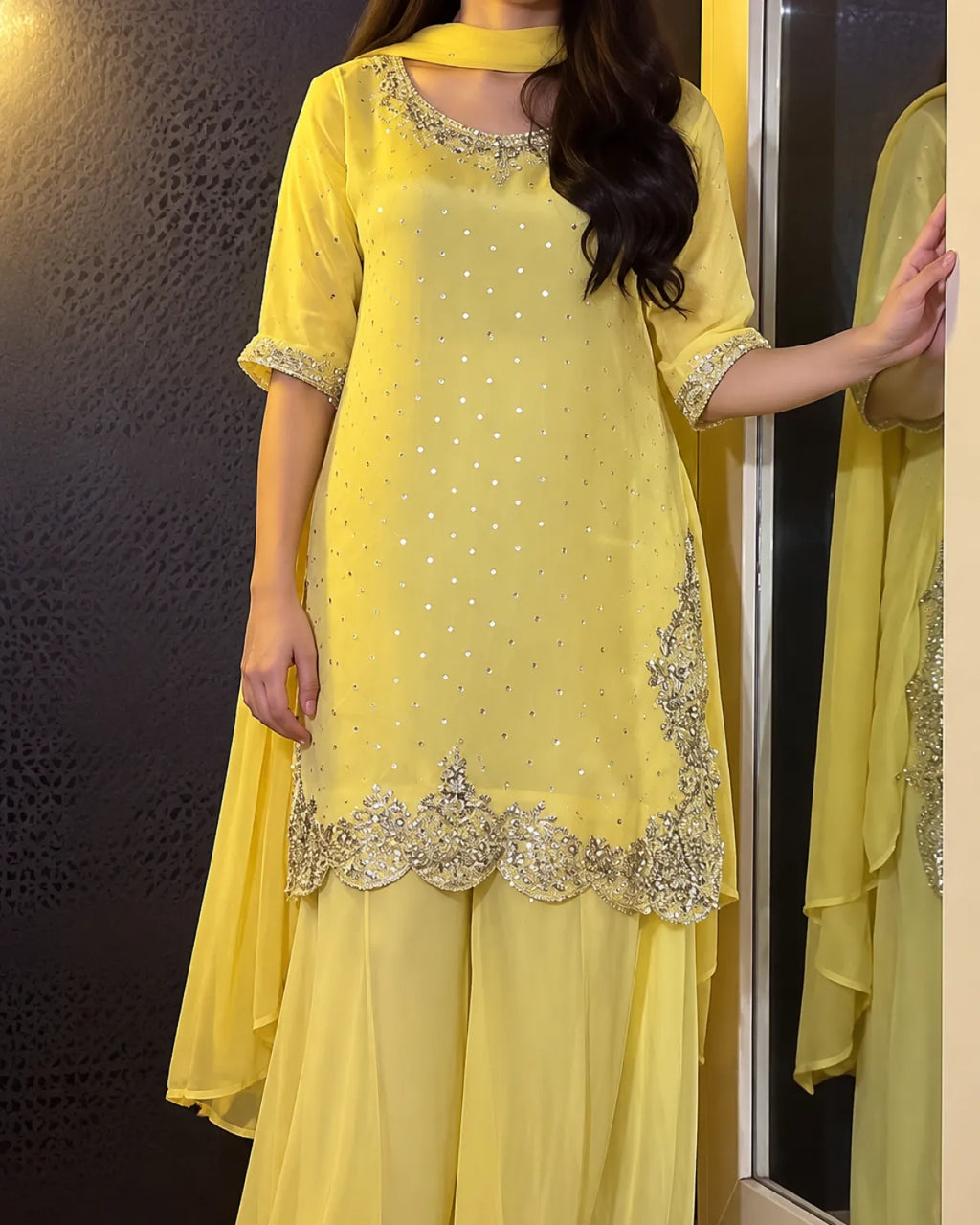 A woman wearing a yellow traditional outfit with silver embroidery and mirror work, standing indoors next to a mirror.