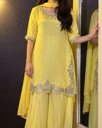 A woman wearing a yellow traditional outfit with silver embroidery and mirror work, standing indoors next to a mirror.