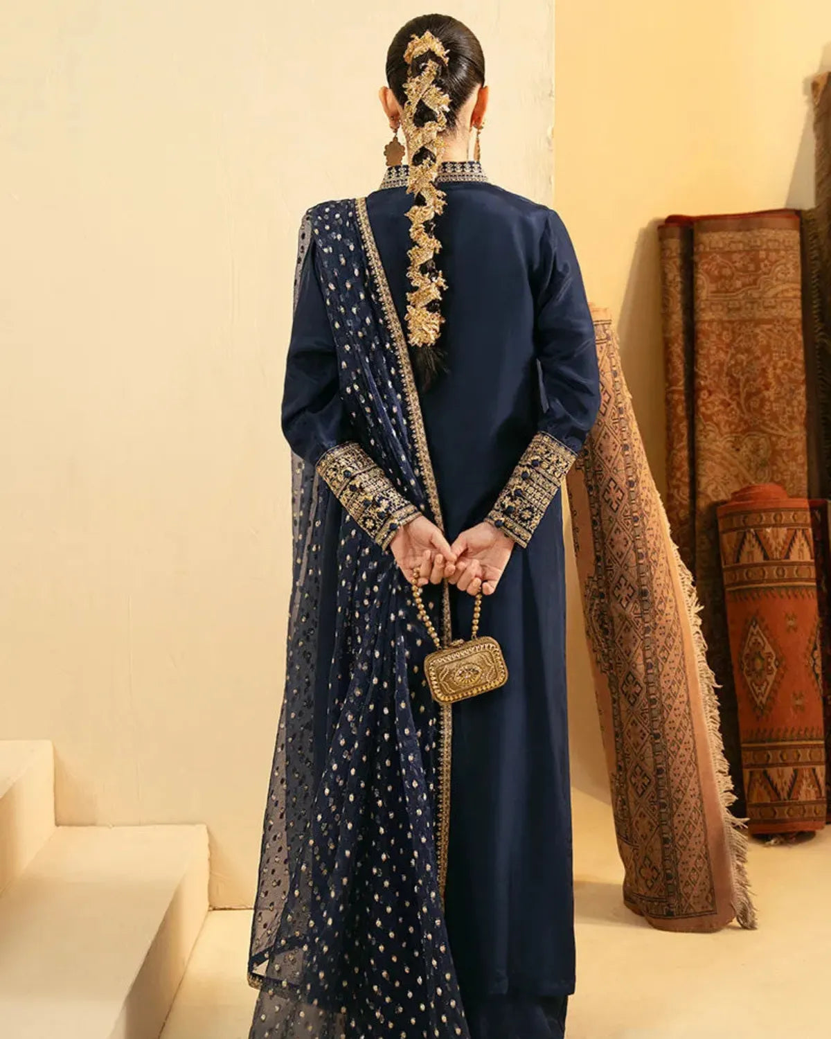 A woman shown from behind wearing a navy blue traditional outfit with gold embroidery and a matching sheer dupatta, standing in a warm beige interior with folded patterned textiles in the background.