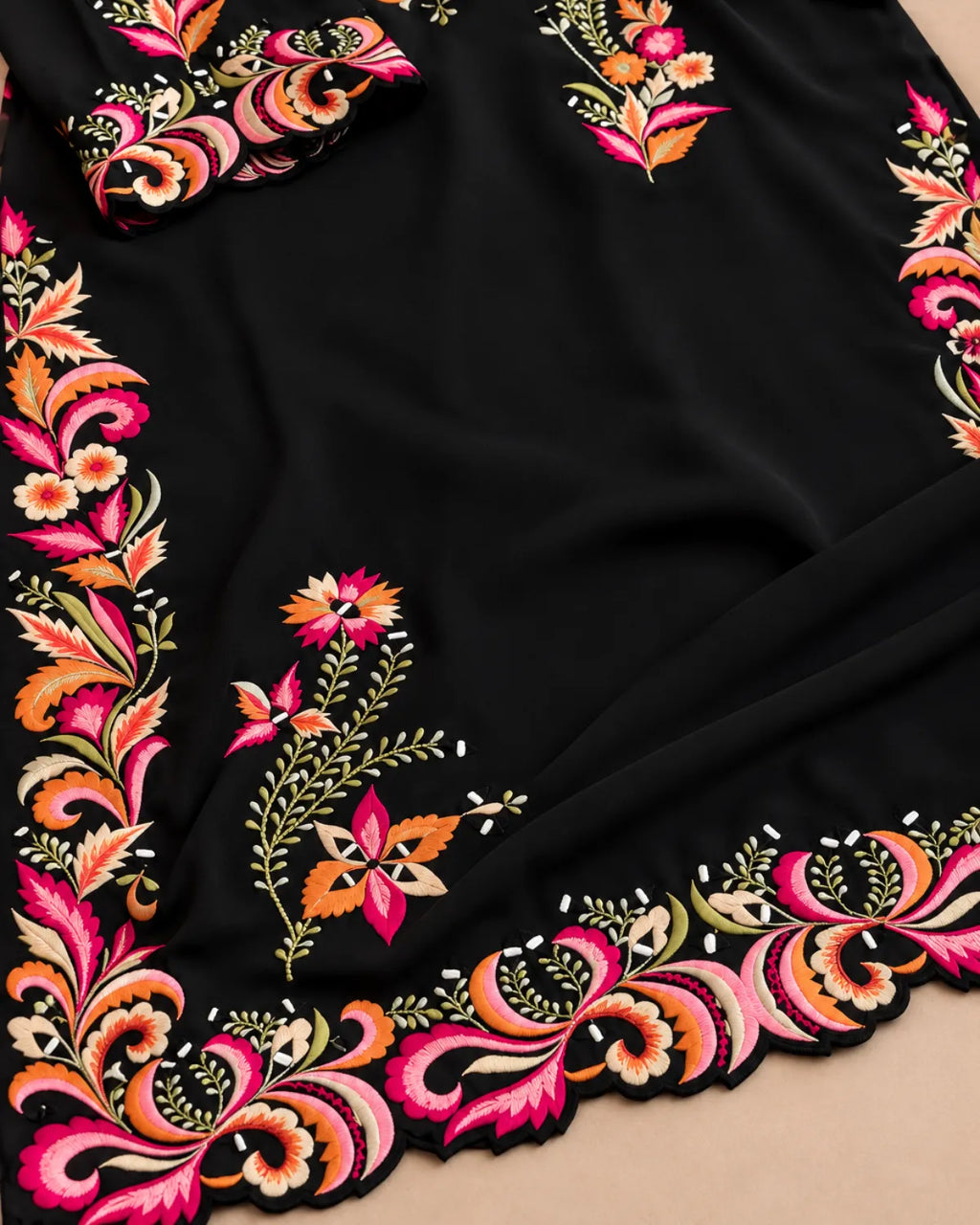 A close-up of a black fabric garment or textile featuring ornate multicolor floral and paisley embroidery along the border, with a matching embroidered cuff or sleeve detail visible at the top left.