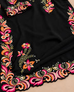 A close-up of a black fabric garment or textile featuring ornate multicolor floral and paisley embroidery along the border, with a matching embroidered cuff or sleeve detail visible at the top left.