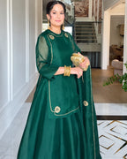 A woman wearing a dark green traditional outfit with gold embroidery, holding a matching green clutch bag with gold embellishments, standing in a modern indoor hallway.