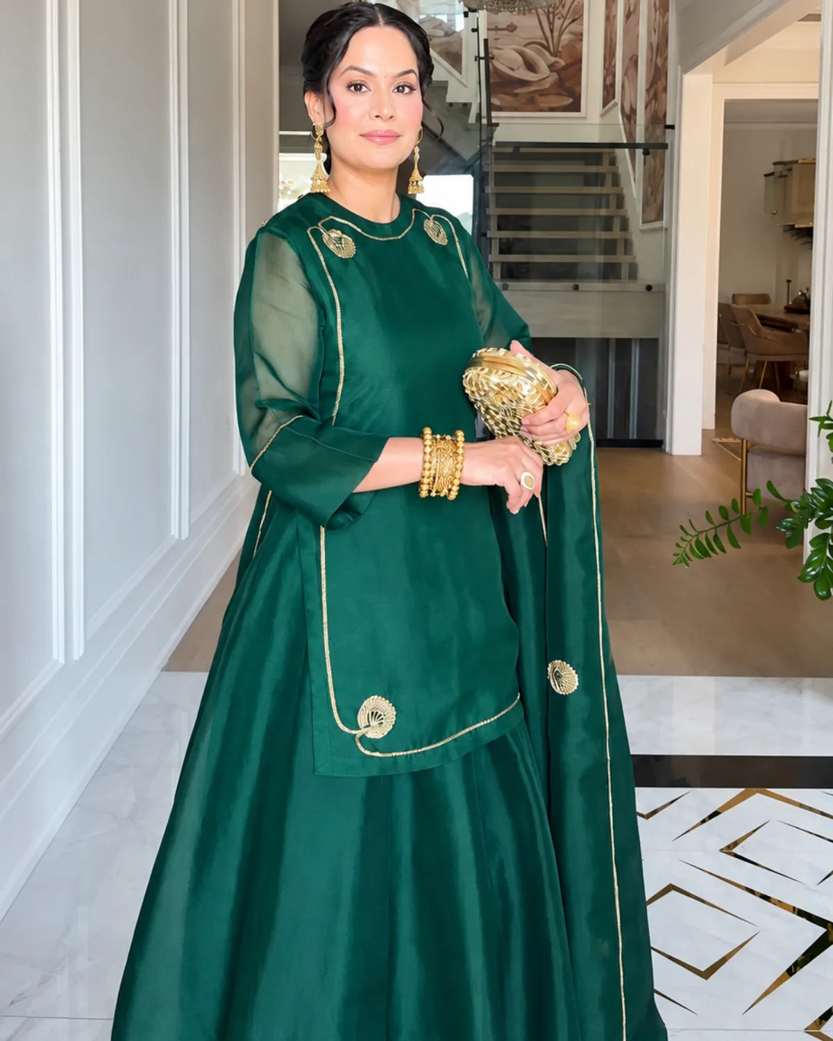 A woman wearing a dark green traditional outfit with gold embroidery, holding a matching green clutch bag with gold embellishments, standing in a modern indoor hallway.