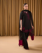 A woman models a black three-piece traditional outfit with deep magenta embroidery and a matching sheer black dupatta, set against a backdrop of gold curtains.