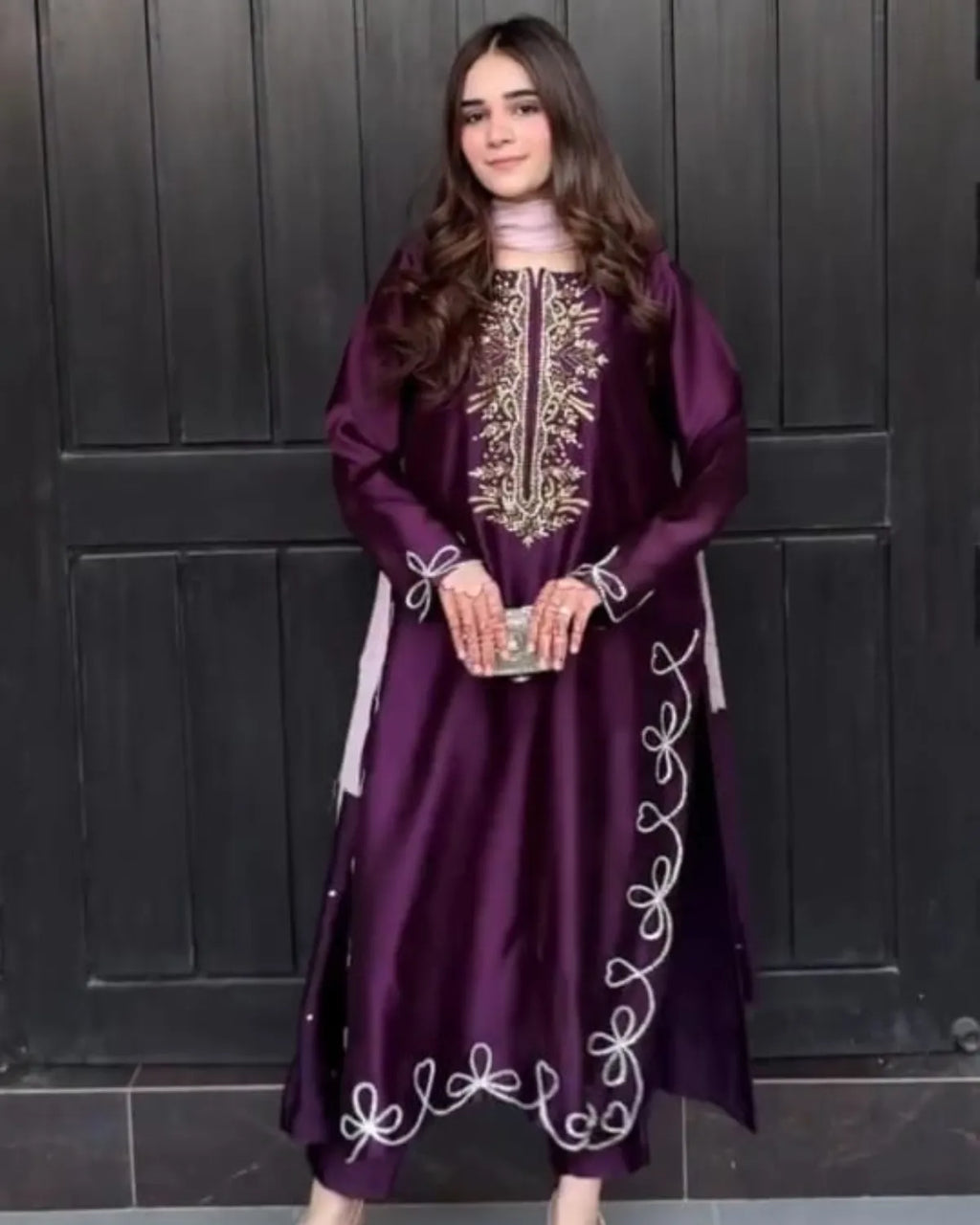 A woman wearing a long, deep purple embroidered dress with white and gold detailing, standing in front of a dark paneled door.