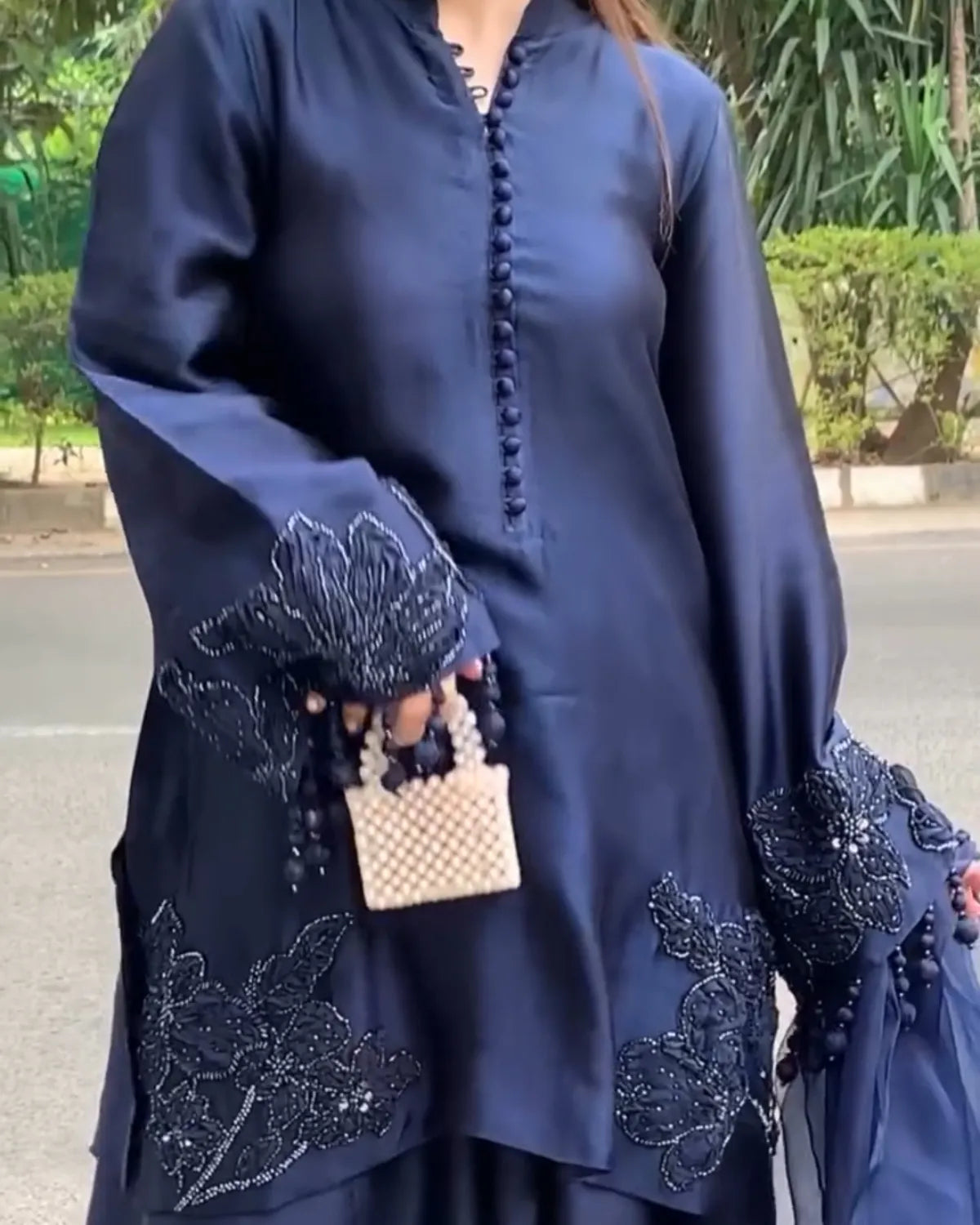 A navy blue satin-like tunic dress with wide bell sleeves, featuring black floral embroidery and beadwork on the cuffs and lower hem, styled with a small woven handbag.
