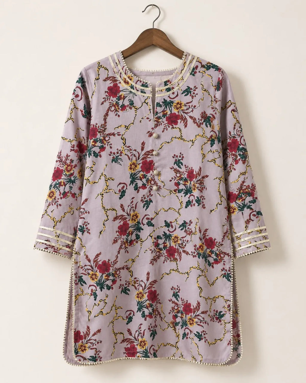 A lavender floral tunic top with gold chain-like embroidery and red, green, and yellow floral motifs, hanging on a wooden hanger against a plain off-white wall.