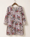 A lavender floral tunic top with gold chain-like embroidery and red, green, and yellow floral motifs, hanging on a wooden hanger against a plain off-white wall.