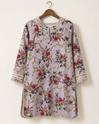 A lavender floral tunic top with gold chain-like embroidery and red, green, and yellow floral motifs, hanging on a wooden hanger against a plain off-white wall.