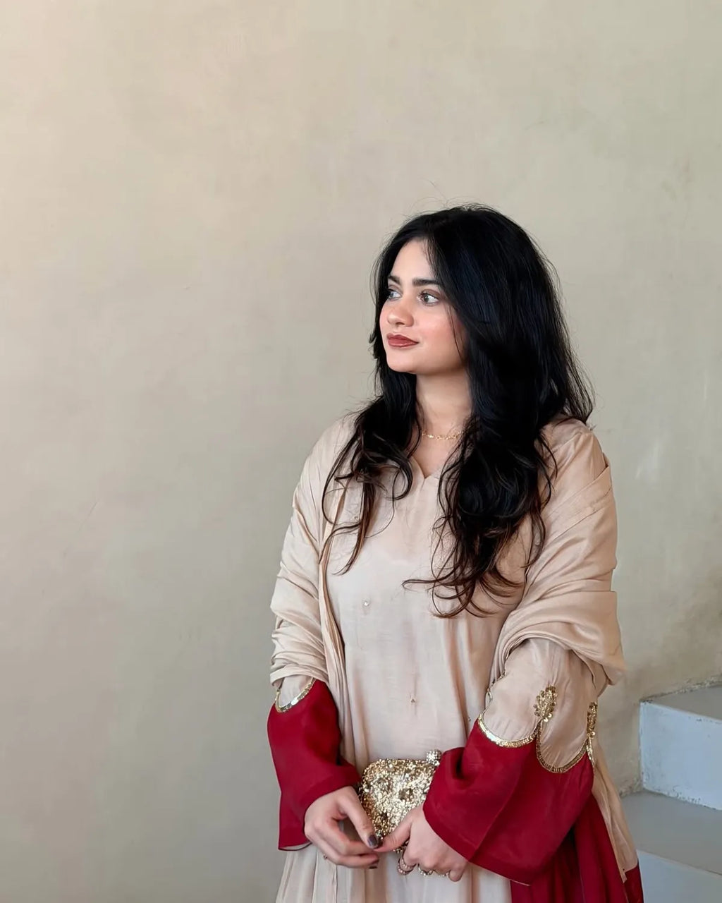 A woman with long black hair is wearing a beige and deep red traditional outfit with gold embroidery, standing against a plain beige wall.