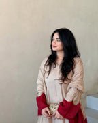 A woman with long black hair is wearing a beige and deep red traditional outfit with gold embroidery, standing against a plain beige wall.