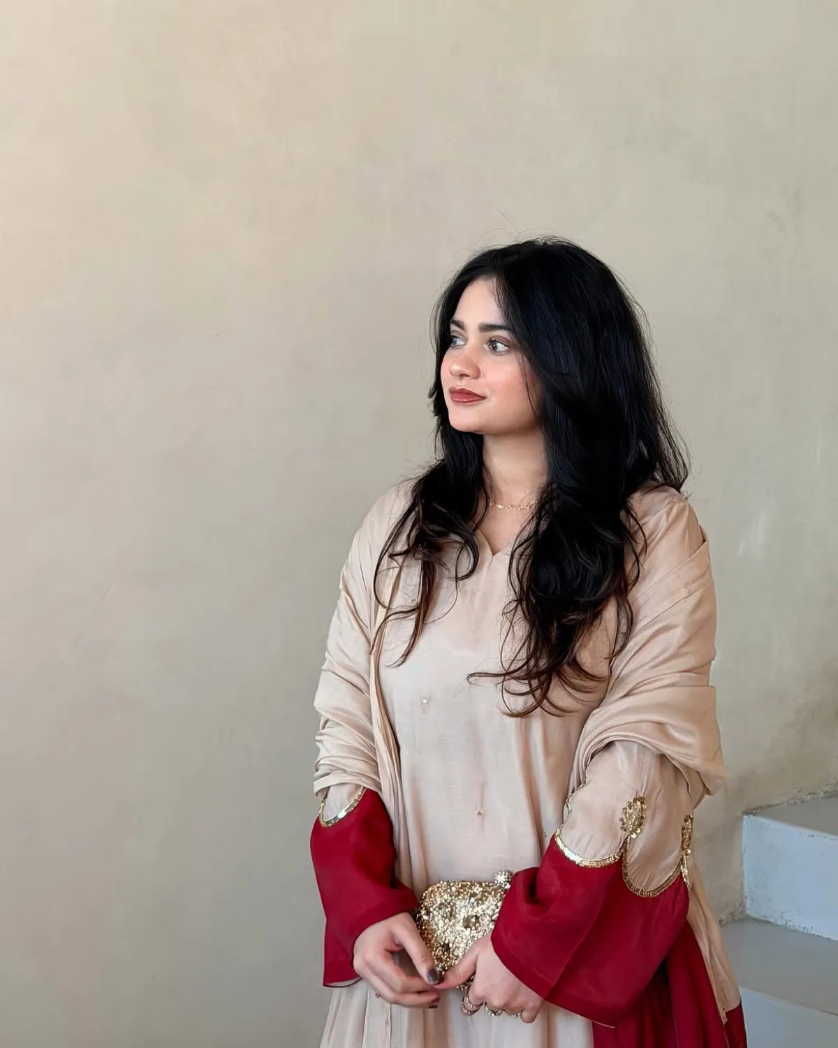 A woman with long black hair is wearing a beige and deep red traditional outfit with gold embroidery, standing against a plain beige wall.