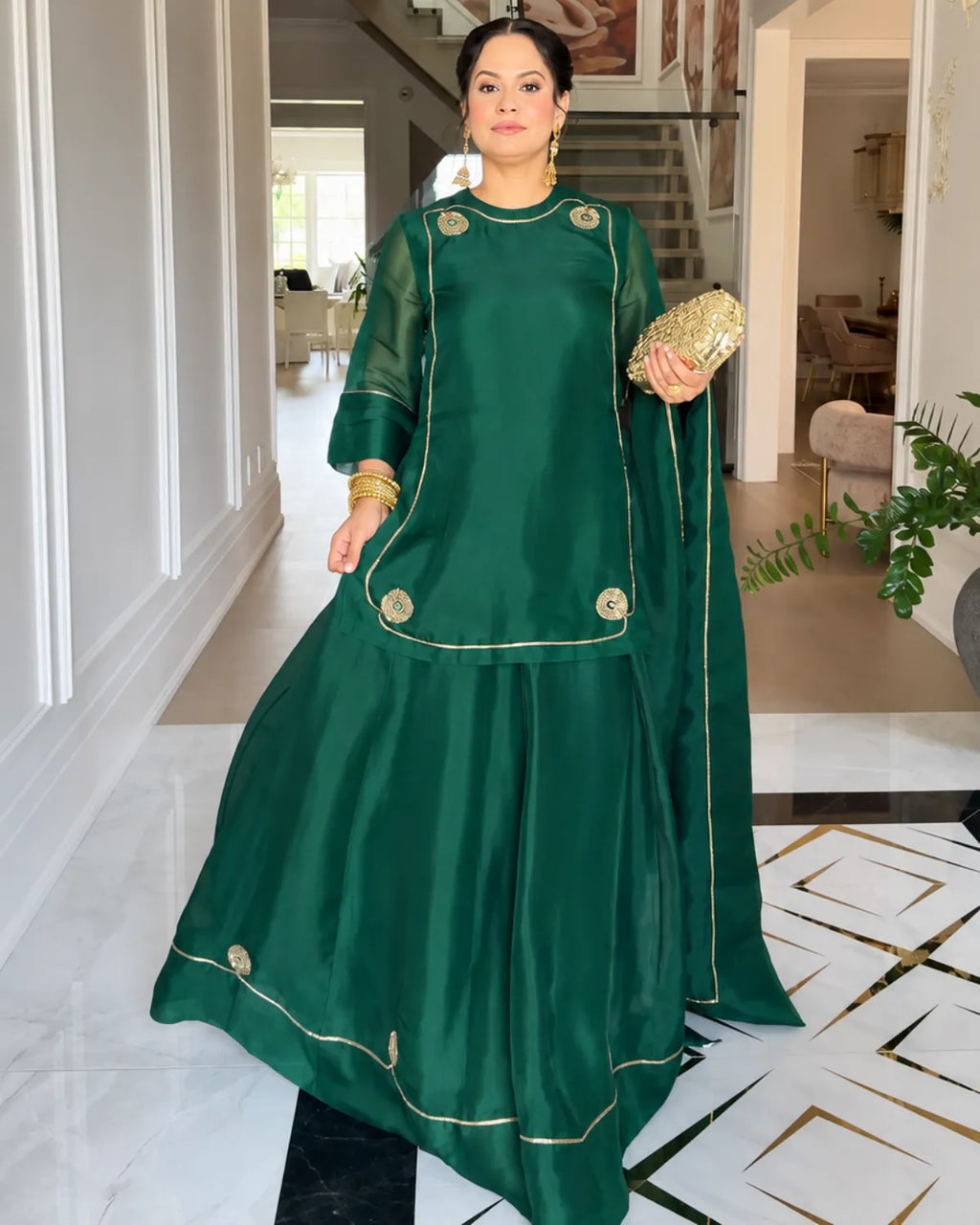 A woman models a dark green traditional outfit with gold embroidery and a matching dupatta in an elegant indoor hallway.