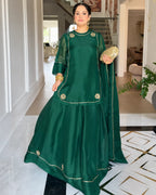 A woman models a dark green traditional outfit with gold embroidery and a matching dupatta in an elegant indoor hallway.