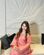 A woman wearing a pink traditional long-sleeve dress with silver trim sits on a beige sofa with black pillows in front of a white decorative wall.