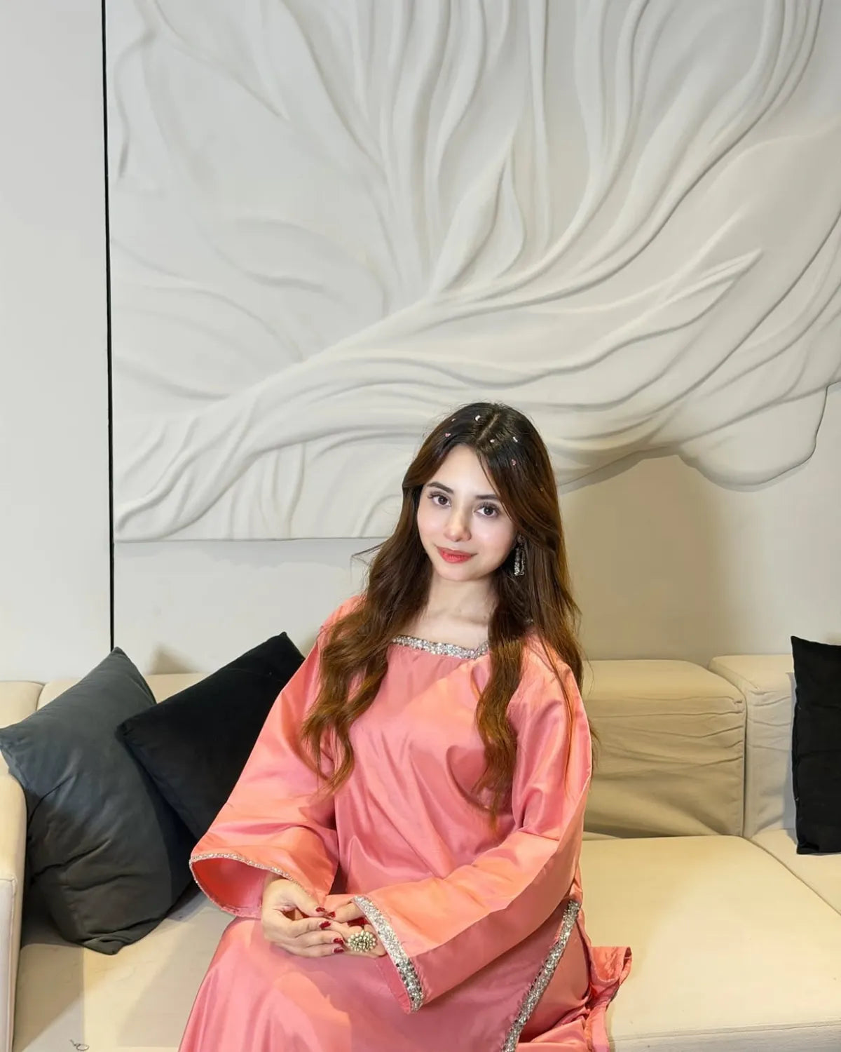 A woman wearing a pink traditional long-sleeve dress with silver trim sits on a beige sofa with black pillows in front of a white decorative wall.
