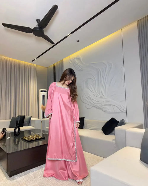 A woman wearing a long pink kaftan dress with silver trim stands in a modern living room with white sofas, a glass coffee table, and a decorative wall.