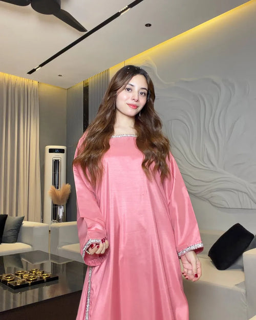 A woman wearing a long-sleeved pink kaftan dress with silver trim, standing in a modern living room setting.