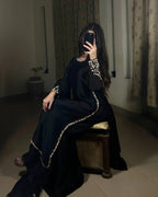 A woman takes a mirror selfie wearing a long black abaya with intricate gold embroidery on the sleeves and along the front hem, sitting on a wooden stool in a dimly lit room.