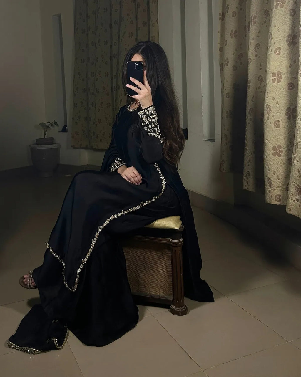 A woman wearing a black traditional outfit with silver embellished trim sits on a wooden stool in a dimly lit room with patterned curtains.