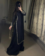 A woman takes a mirror selfie wearing a long black dress with ornate gold embroidery on the sleeves and along the front drape. She is standing in an indoor room with patterned curtains and tiled flooring.
