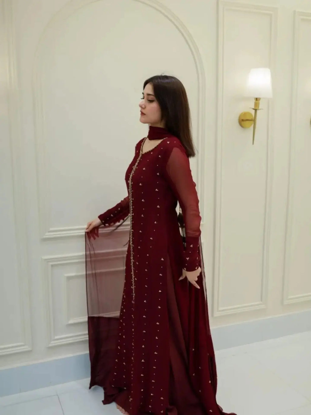A woman wearing a deep maroon long gown with silver embellishments, standing in a white paneled room.