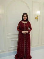 A woman wearing a long maroon red embroidered gown with sheer sleeves and a high neckline, standing in front of a white paneled wall with a wall lamp.